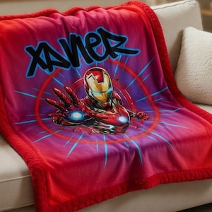 Puede incluir: Manta de felpa roja y rosa con un gr&aacute;fico de Iron Man y el nombre "Xavier" en letras azules estilo grafiti. La manta tiene un borde rojo y est&aacute; sobre un sof&aacute; blanco.