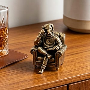 Puede incluir: Figura de bronce de un perro sentado en un sillón pequeño, leyendo un libro. El perro lleva un suéter y parece estar pensativo. La figura está colocada sobre una superficie de madera, con un vaso de líquido ámbar y un posavasos cerca.