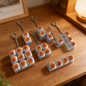 May include: A collection of keychains featuring a design of small, square, orange and white blocks. The keychains have silver-colored metal hardware. The keychains are arranged on a wooden surface.