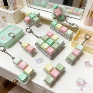 Könnte beinhalten: Eine Sammlung von Schlüsselanhängern mit pastellfarbenen Tastenkappen. Die Tastenkappen sind in Rosa, Grün und Gelb gehalten und in verschiedenen Konfigurationen angeordnet. Jeder Schlüsselanhänger hat einen silberfarbenen Ring. Die Artikel werden auf einer weißen Oberfläche präsentiert.