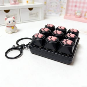 Könnte beinhalten: Schwarzer Schlüsselanhänger mit einem Neun-Tasten-Tastaturdesign, jede Taste mit einem rosa Pfotenabdruck versehen. Die Tasten sind in einem 3x3-Raster angeordnet. Ein schwarzer Verschluss ist angebracht. Eine kleine weiße Katzenfigur ist im Hintergrund zu sehen.