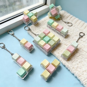 May include: Various pastel-coloured keycap accessories, including keyrings and a square display piece. The keycaps are in shades of pink, yellow, and green, set in clear plastic cases. The keyrings have silver-toned metal rings.