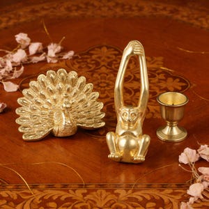 May include: A collection of gold-colored decorative items, including a peacock figurine with a fanned tail, a monkey figurine with arms raised, and a small candle holder. The items are arranged on a dark wooden surface.
