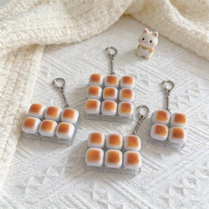 May include: Four keychains featuring miniature, square-shaped food items with a light brown top and white base, arranged in a grid. Each keychain has a silver-colored ring. A small, white bear figurine with orange accents is also present.