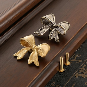 May include: Two decorative bow-shaped cabinet knobs in antique gold and bronze tones. The knobs are made of metal and have detailed textures. Two matching screws are included, ready for installation. The knobs are set on a dark brown wooden surface.