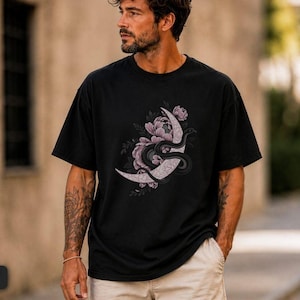 May include: A black short-sleeved t-shirt featuring a graphic print on the chest. The design displays a crescent moon decorated with light purple flowers and a dark, winding snake coiled around the moon's surface, set against a solid black fabric.