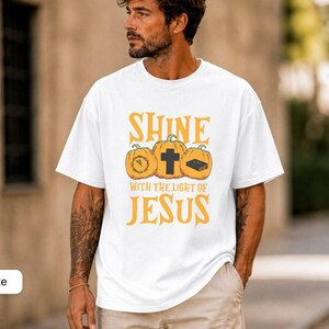 May include: A white short-sleeved t-shirt featuring a graphic with three orange pumpkins. The centre pumpkin has a black cross, the left has a clock, and the right has a book. The text reads SHINE WITH THE LIGHT OF JESUS in yellow.