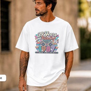 May include: A white short-sleeve t-shirt featuring a colourful graphic print that includes a boombox, musical notes, and the text 39 Years of Retro Cool 1987. The design uses a palette of pink, blue, and yellow with geometric shapes.