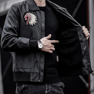 Op de afbeelding: Zwarte imitatie leren jas met een klassieke kraag, een ritssluiting en een Native American chief patch op de borst. De jas heeft een zwarte geribbelde zoom en manchetten en twee voorzakken. Een horloge is zichtbaar op de pols.