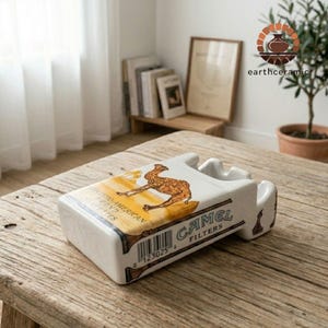 以下が含まれることがあります： キャメルフィルターのタバコのパッケージを模した長方形の白い陶器製灰皿。上面には砂漠の風景の中にラクダのグラフィック、Camel Filtersの文字、バーコードが描かれており、タバコを置くための溝が一体化されています。