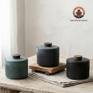 May include: Three ceramic jars with wooden lids. The jars are in shades of green, grey, and black, with vertical ribbed detailing. The jars are displayed on a wooden table with a neutral background. The logo "earthceramic" is visible.