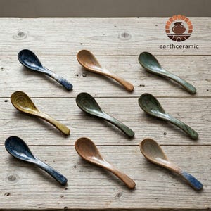May include: Eight ceramic spoons in shades of blue, brown, and green, displayed on a wooden surface. The spoons have a handcrafted look. The logo "earthceramic" is visible. The spoons are suitable for serving food.