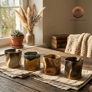 May include: Four handcrafted ceramic cups with a unique, textured design. The cups feature a mix of brown, tan, and black hues, sitting on a neutral-colored linen cloth. The rustic aesthetic is complemented by a wooden table and natural light.
