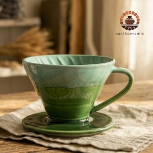 Puede incluir: Un vertedor de café de cerámica de dos tonos, verde y verde azulado, con un platillo a juego. El vertedor tiene un asa y está sobre un paño de color neutro. La marca "earthceramic" es visible.