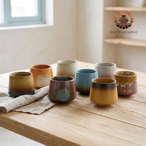 Op de afbeelding: Een verzameling van acht keramische bekers met een rustieke, handgemaakte uitstraling. De bekers hebben verschillende kleuren, waaronder bruin, oranje, blauw en beige, met een gradiënteffect. De merknaam "earthceramic" is zichtbaar.