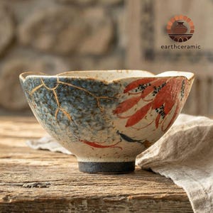 Może przedstawiać: Ceramiczna miska o unikalnym designie. Misa ma niebiesko-białą kolorystykę ze złotymi akcentami i czerwonymi wzorami przypominającymi liście. Misa ma ciemną podstawę i tekst "earthceramic".