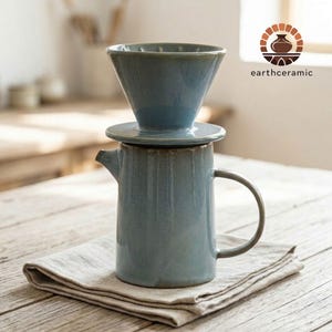 May include: A blue ceramic pour-over coffee maker set. The set includes a cone-shaped dripper, a lid, and a cylindrical carafe with a handle. The set is placed on a folded linen napkin. The logo "earthceramic" is visible in the upper right corner.