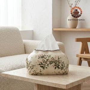 Pode incluir: Uma caixa de lenços de cerâmica na cor creme com formato retangular arredondado. A superfície apresenta uma estampa botânica detalhada em tons de verde e rosa suave. Um lenço branco sai pela abertura superior do recipiente.