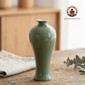 May include: A sage green ceramic vase with a glossy finish. The vase has a slender, elongated shape with a flared base and a narrow neck. The vase is displayed on a wooden table. The logo "earthceramic" is visible in the upper right corner.