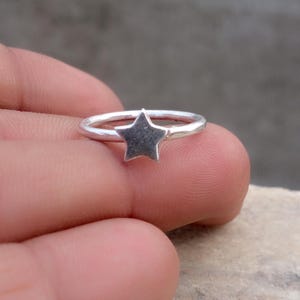 以下が含まれることがあります： 細く滑らかな円形のバンドの中央に、小さな五芒星のチャームがあしらわれたシルバーカラーのリング。金属の表面は磨き上げられた仕上げで、星はバンドの前面にしっかりと固定されており、シンプルで統一感のあるデザインとなっています。