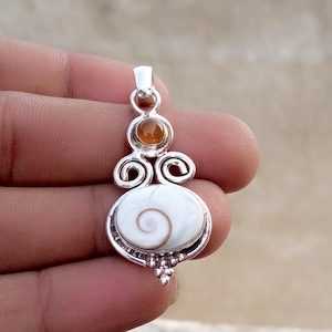 Puede incluir: Un colgante de plata con una gran concha de ojo de shiva ovalada y blanca con un patrón de espiral natural, acentuado por una piedra redonda más pequeña de color ámbar encima. El engaste metálico incluye espirales de plata y cuentas.
