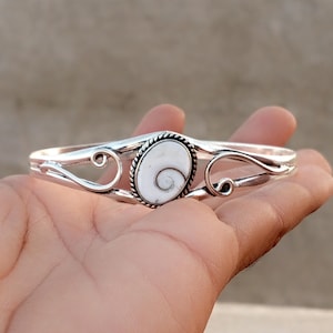 May include: A silver cuff bracelet featuring a central oval-shaped white shiva eye shell with a dark spiral pattern. The metal band is decorated with twisted rope detailing around the stone and symmetrical swirling scrollwork on both sides.