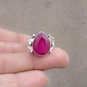 May include: A silver ring featuring a large, pear-shaped, faceted deep pink ruby gemstone set in a metal frame. The band consists of multiple thin silver wires that converge at the sides, accented with small, polished silver spheres on each side.