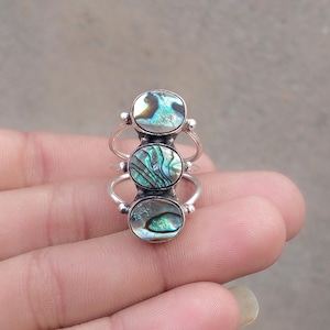May include: A silver ring featuring three vertically aligned oval abalone shell cabochons. The iridescent shell displays swirling patterns of green, blue, and brown. The metal band is split into three thin, polished silver wires that connect to the settings.