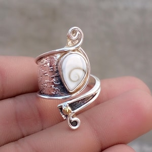 May include: A silver bypass ring featuring a teardrop-shaped white shiva eye shell with a spiral pattern. The band has a textured, oxidized finish with a polished silver swirl at the top and a small gold-toned bead accent at the bottom terminal.