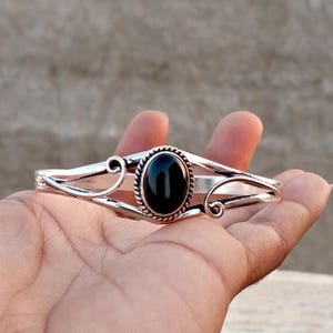 May include: A silver-toned cuff bracelet featuring a central oval-shaped black stone set within a twisted rope-style border. The metal band extends outwards with decorative swirling scrollwork on both sides of the prominent black gemstone.