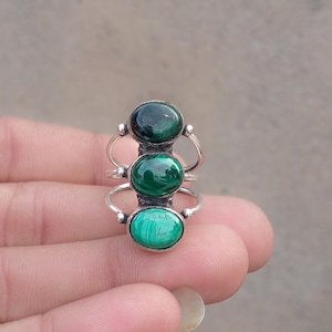 May include: A silver ring featuring three vertically aligned oval malachite gemstones. Each stone displays distinct green banding patterns. The stones are set in polished silver bezels, connected by a multi-band silver wire shank design.