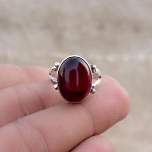 May include: A silver ring featuring a large, oval-shaped, deep red gemstone set in a smooth bezel. The band of the ring has a split design with small, decorative silver spheres accenting the sides where the band meets the central stone setting.