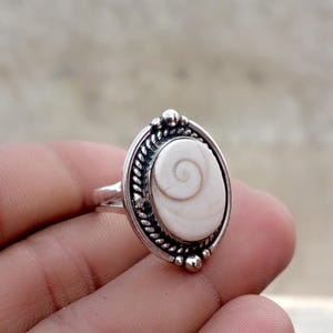 Pode incluir: Um anel de prata com uma incrustação oval de concha de olho de Shiva branca com um padrão espiral natural. A cravação é emoldurada por uma borda de metal trançado e pequenas esferas de prata no topo e na base do centro oval.