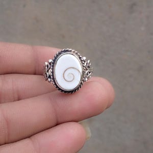 May include: A silver ring featuring an oval-shaped white stone with a natural brown spiral pattern. The setting is crafted with a twisted rope-like border and intricate metal scrollwork details along the sides of the band, creating a detailed look.