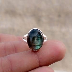 Puede incluir: Un anillo de plata con una gran piedra preciosa de labradorita de forma ovalada. La piedra muestra una superficie oscura e iridiscente con destellos azules y verdes visibles. La banda de metal tiene un diseño dividido que se une al engaste.