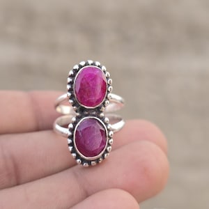 Puede incluir: Un anillo de plata con dos piedras preciosas de rubí de color rosa intenso, de forma ovalada y facetadas, apiladas verticalmente. Cada piedra está engastada en un bisel de plata con cuentas decorativas y el aro es de dos hilos.