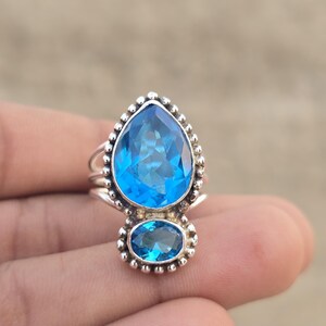Puede incluir: Un anillo de plata de ley con una gran piedra azul en forma de pera sobre una piedra azul más pequeña de forma ovalada. Ambas piedras están rodeadas por un borde de metal con cuentas y la banda del anillo consta de tres hilos de plata finos.