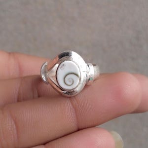 Pode incluir: Um anel de tom prateado com uma pedra central oval branca que apresenta um padrão de espiral castanho-escuro. A banda de metal tem um acabamento liso e polido, com uma cravação larga que emoldura a pedra incrustada para um visual limpo.