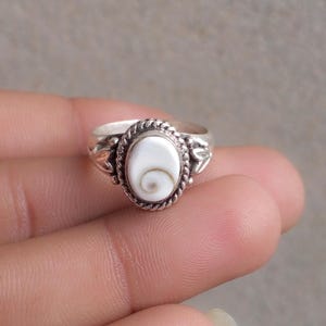 Op de afbeelding: Een zilveren ring met een ovaalvormige Shiva-oog schelp inlay. De witte schelp in het midden vertoont een natuurlijk bruin spiraalpatroon. De zetting is omlijst door een gedraaide touwrand en decoratieve zilveren bladachtige accenten.