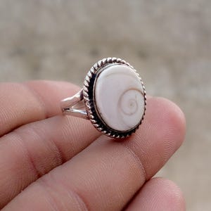 May include: A silver ring featuring an oval-shaped shiva eye shell inlay. The centre of the ring displays a natural spiral pattern in shades of white and cream. The setting is framed by a twisted rope-style metal border on a split band.