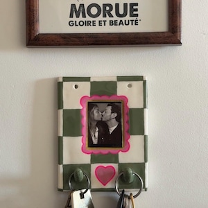 Pode incluir: Ilustração em preto e branco emoldurada de um peixe com o texto «MORUE GLOIRE ET BEAUTÉ» acima de um porta-chaves. O porta-chaves tem uma foto emoldurada em rosa, um padrão xadrez verde e branco e um coração rosa.
