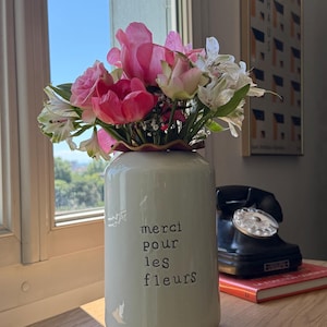 Peut inclure: Un vase en céramique blanc rempli de fleurs roses et blanches, avec l'inscription "merci pour les fleurs" en noir. Le vase est posé sur une surface en bois près d'une fenêtre, avec un téléphone noir vintage et un livre en arrière-plan.