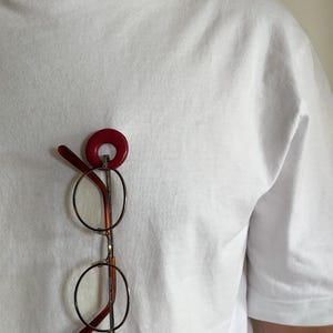 May include: A plain white short-sleeved t-shirt featuring a pair of round-framed glasses with dark red temples suspended from a circular, deep red accessory attached to the fabric. The glasses have thin metallic rims and clear lenses.