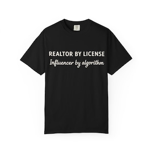 Puede incluir: Camiseta negra con texto blanco. El texto dice "REALTOR BY LICENSE" encima de "Influencer by algorithm". La camiseta tiene cuello redondo y mangas cortas. El texto está centrado en la parte delantera.