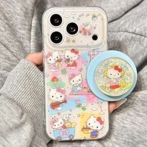 May include: Clear phone case with a colorful Hello Kitty design. The case features multiple illustrations of Hello Kitty in various scenes, along with a light blue pop socket with a Hello Kitty design. The case is held by a person.