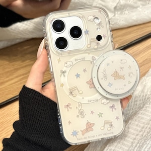 Könnte beinhalten: Transparente Handyhülle mit cremefarbenem Hintergrund, verziert mit Noten, Sternen, Schleifen und einer Hundeillustration. Ein passender Pop-Socket ist angebracht. Der Text "Each day brings a new beginning" ist aufgedruckt.