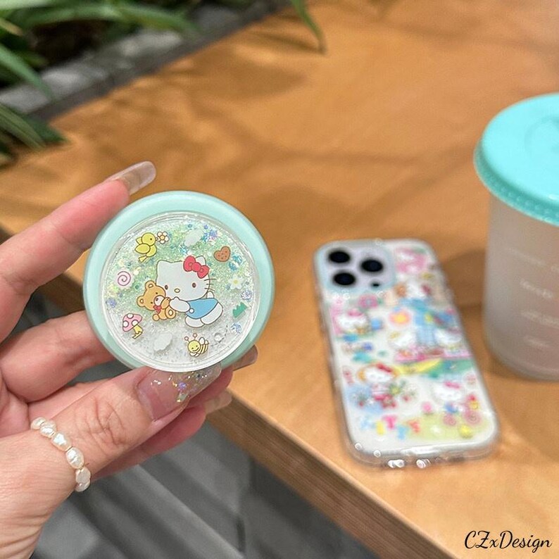 Pu&ograve; includere: Un popsocket azzurro con Hello Kitty e amici, con uno sfondo trasparente e glitterato. &Egrave; visibile anche una custodia trasparente per telefono con un design di Hello Kitty. Il popsocket &egrave; tenuto da una persona.