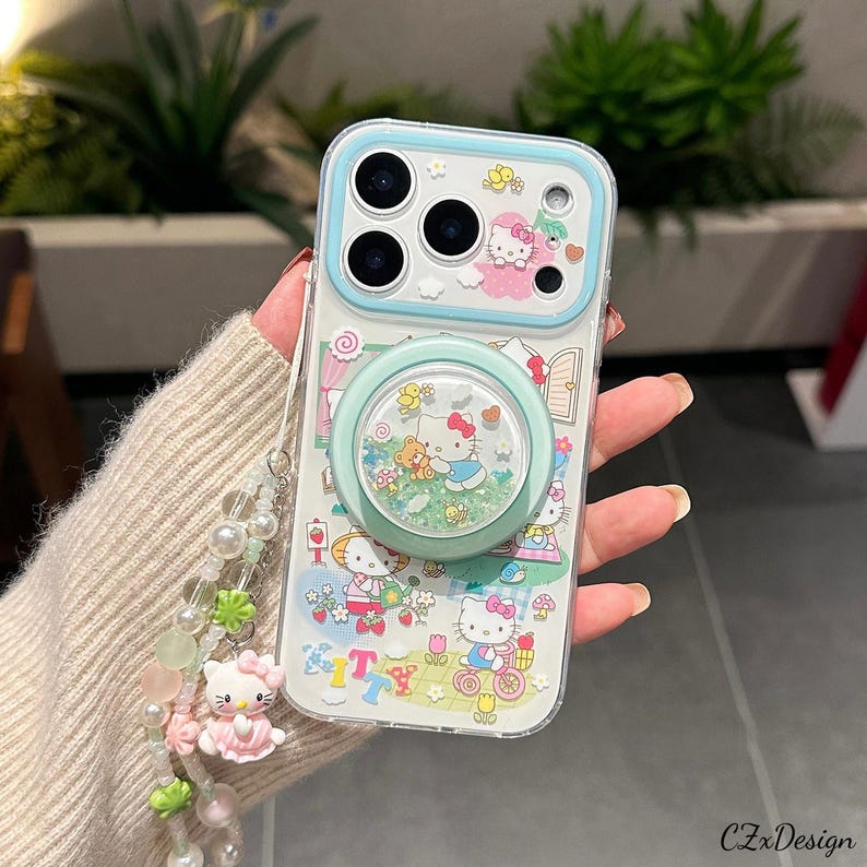 Pu&ograve; includere: Custodia trasparente per telefono con design Hello Kitty, bordo azzurro e supporto abbinato. La custodia presenta un'illustrazione a cartoni animati con personaggi di Hello Kitty, fiori e altri elementi decorativi. &Egrave; attaccato un ciondolo di perline.