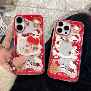 May include: Two clear phone cases with red borders and Hello Kitty designs. One case features a large Hello Kitty face with a red bow and floating confetti. The other case has multiple Hello Kitty characters, snowmen, and gingerbread men.