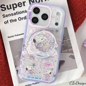 Puede incluir: Funda transparente para teléfono con diseño de Snoopy. La funda tiene un borde lila y presenta ilustraciones de Snoopy con estrellas, corazones y el texto "GOOD LUCK EVERY DAY". También tiene un agarre emergente.